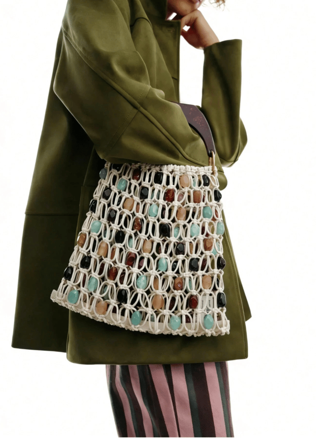 A person wearing an olive green coat and pink striped trousers holds a unique beaded bag with multi-coloured stones or beads, against a plain white background.