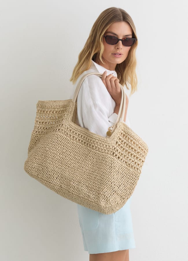 Woman with blonde hair wearing sunglasses, a white shirt, and light blue shorts stands against a white background, holding a large woven basket bag over her shoulder.