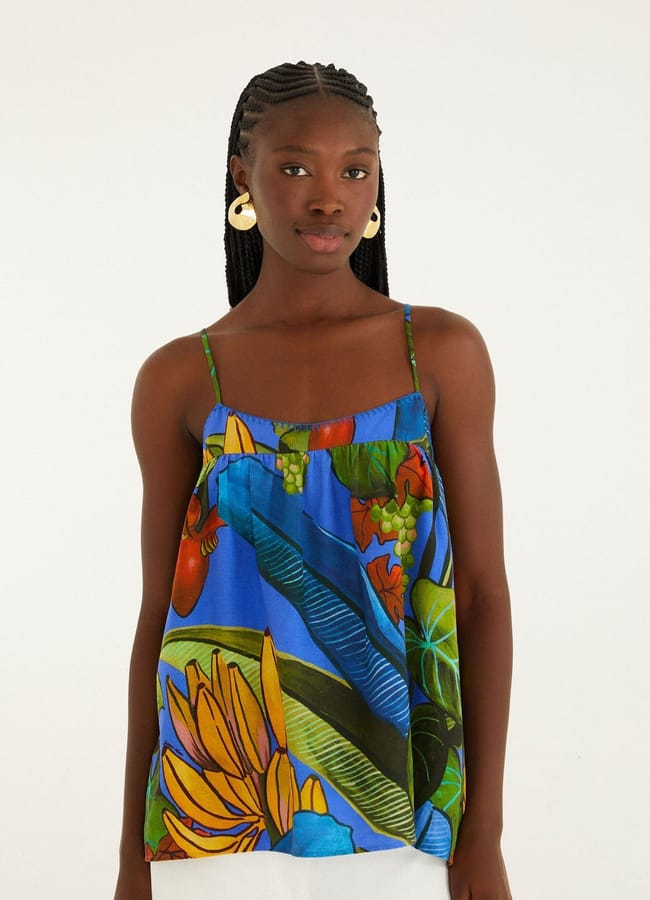 A woman with plaited hair and large hoop earrings wears a colourful, blue camisole with thin straps featuring tropical fruit prints and white trousers, standing against a plain, light background.