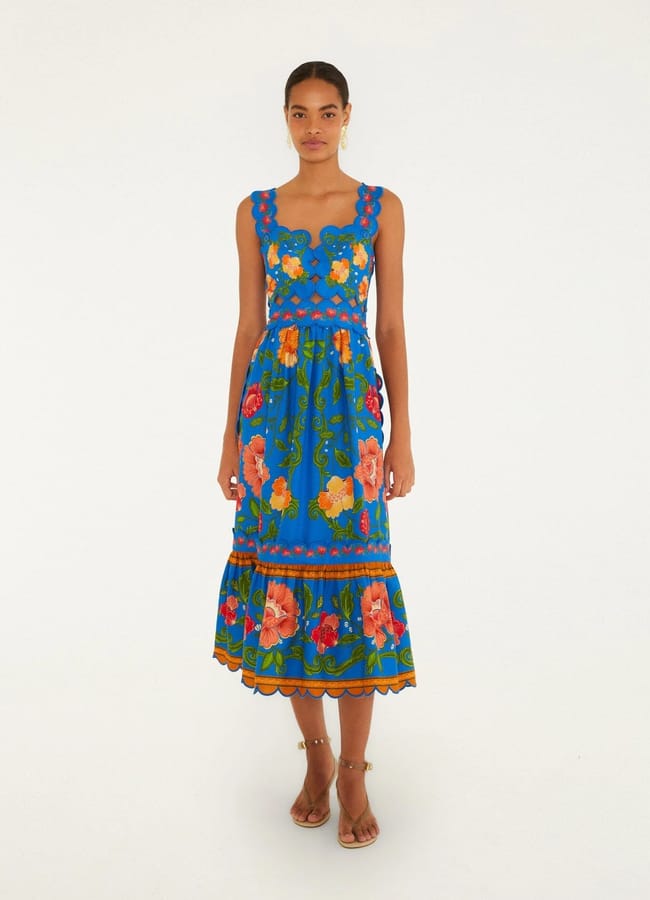 A woman stands against a plain background wearing a sleeveless, blue midi dress with a scalloped hem and colourful floral patterns. She has her hair tied back and is wearing strappy sandals.