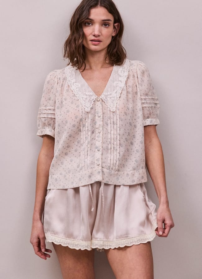 A woman stands against a plain background, wearing a light, sheer summer top with a lace collar and pleated front, paired with silky, light-coloured shorts featuring lace trim at the hem.