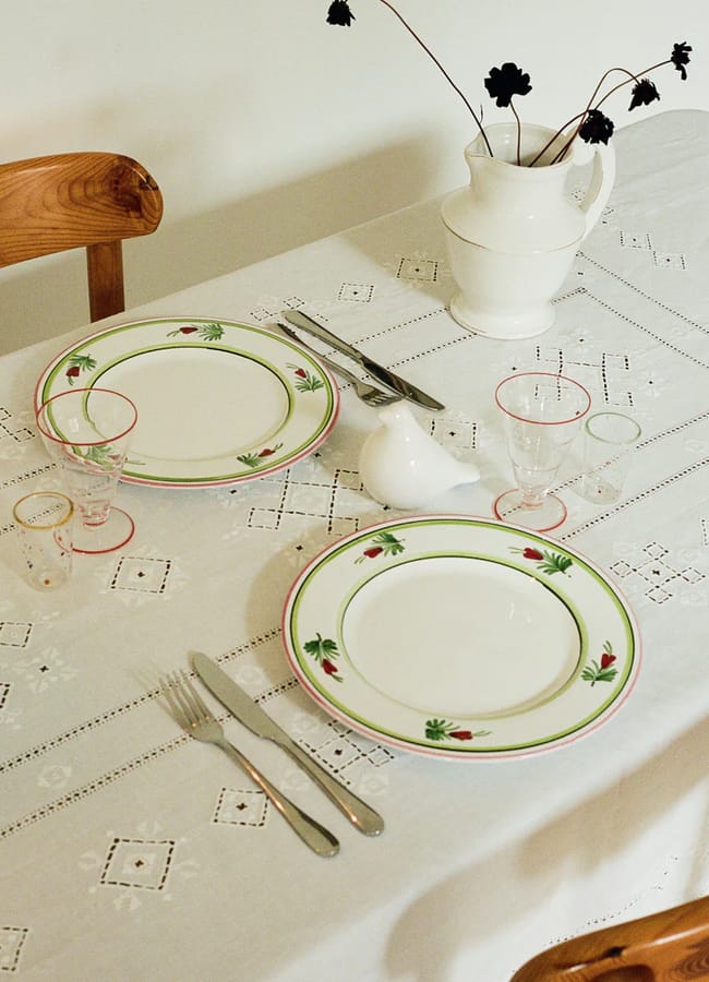 A neatly set dining table with a white embroidered tablecloth, two plates with green and red rims, silver cutlery, clear glasses, and a white jug with black flowers. A wooden chair is beside the table.