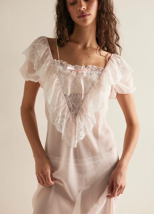 A woman wearing a sheer, light pink nightdress with lace frill details and a small bow at the neckline stands against a plain light background—an elegant idea for Father’s Day gifts 2026.