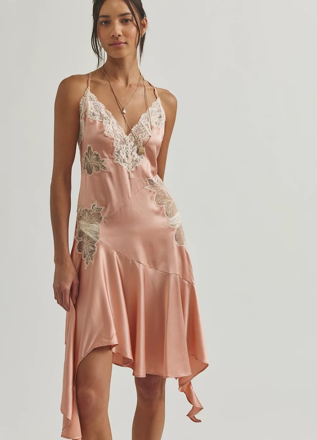 A woman wearing a light pink satin slip dress with white lace and floral appliqué details, thin straps, and an asymmetrical hem stands against a plain light background—perfect inspiration for unique Fathers’ Day gifts 2026.