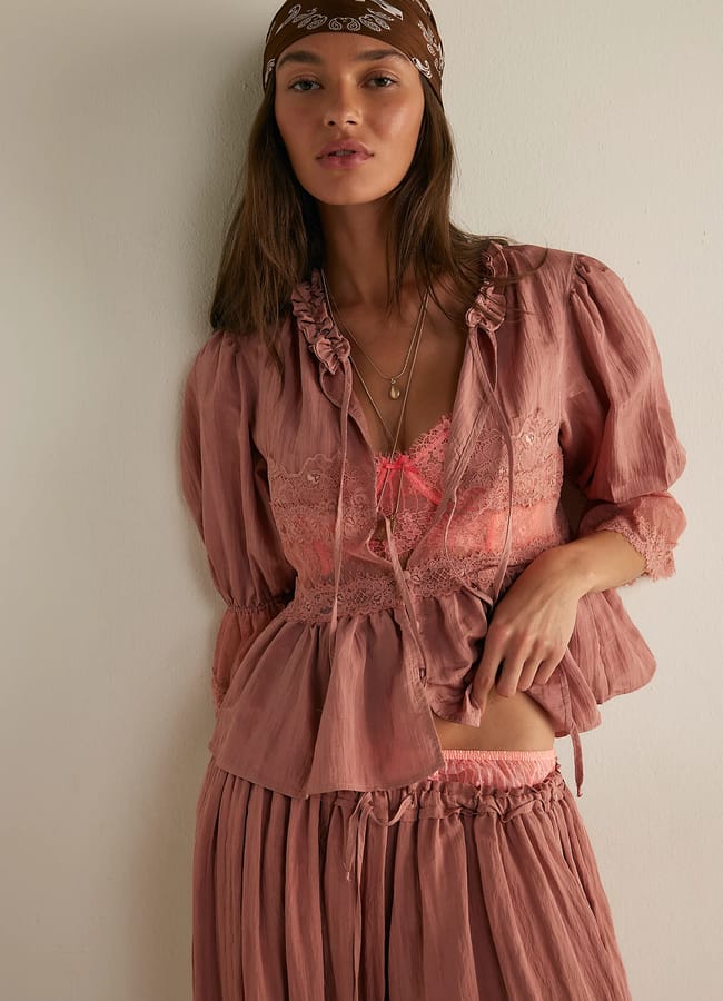 A woman wearing a loose, dusty rose-coloured summer top and matching skirt, a pink lace bralette, layered necklaces, and a brown patterned bandana stands against a light wall, gazing at the camera.