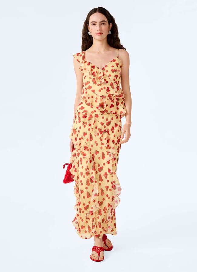 A woman wearing a sleeveless, frilled, cream-coloured dress with red fruit prints, red sandals, and holding a small red bag stands against a plain light background.