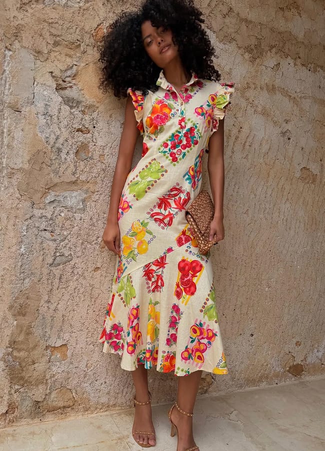 A woman with curly hair poses against a textured stone wall, wearing a cream-coloured, ruffled sleeveless dress adorned with colourful floral and fruit prints. She holds a woven clutch and completes the look with strappy sandals.