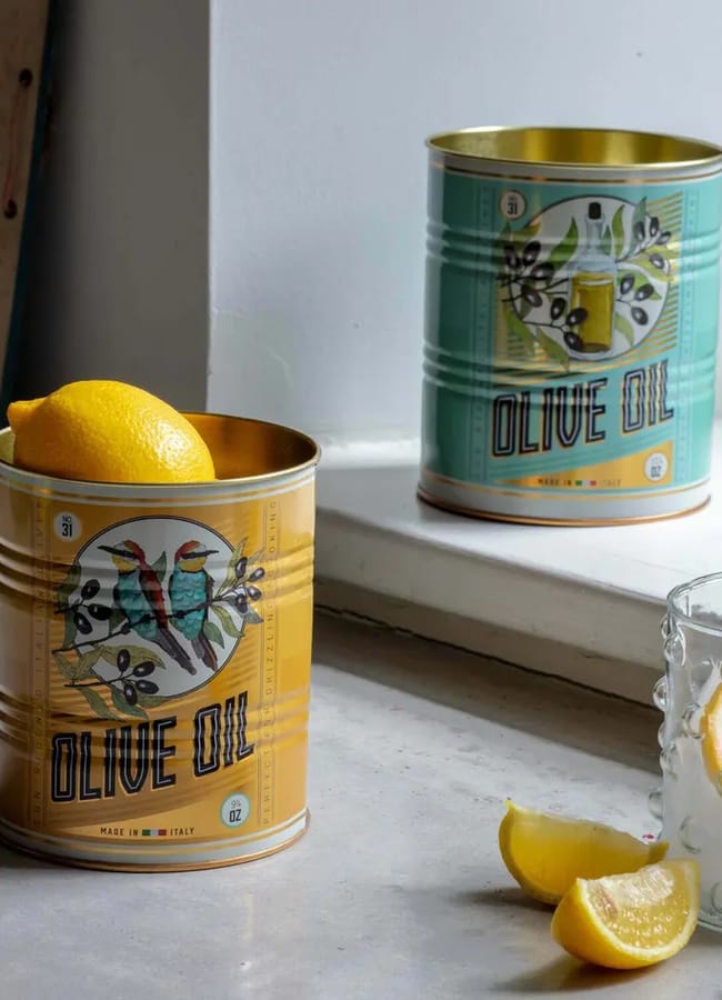 A yellow olive oil tin holding a lemon sits on a counter next to a textured glass of water with lemon. A green olive oil tin is in the background, reflected in a window.