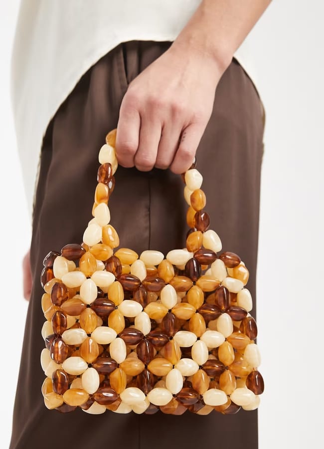 A person holds a beaded bag, its small rectangular shape crafted from brown, amber, and cream-coloured beads with a matching handle. The look is completed with brown trousers and a light top.