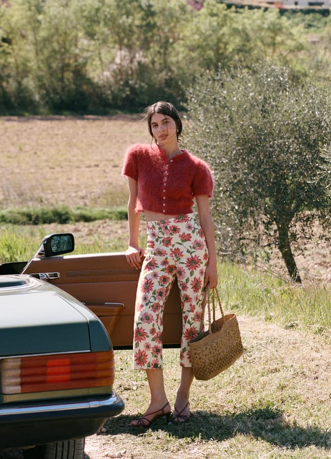 A woman stands outdoors by an open car door, wearing a fuzzy red short-sleeve cardigan and floral print trousers. She holds a woven straw bag and is surrounded by greenery and trees in a rural setting.