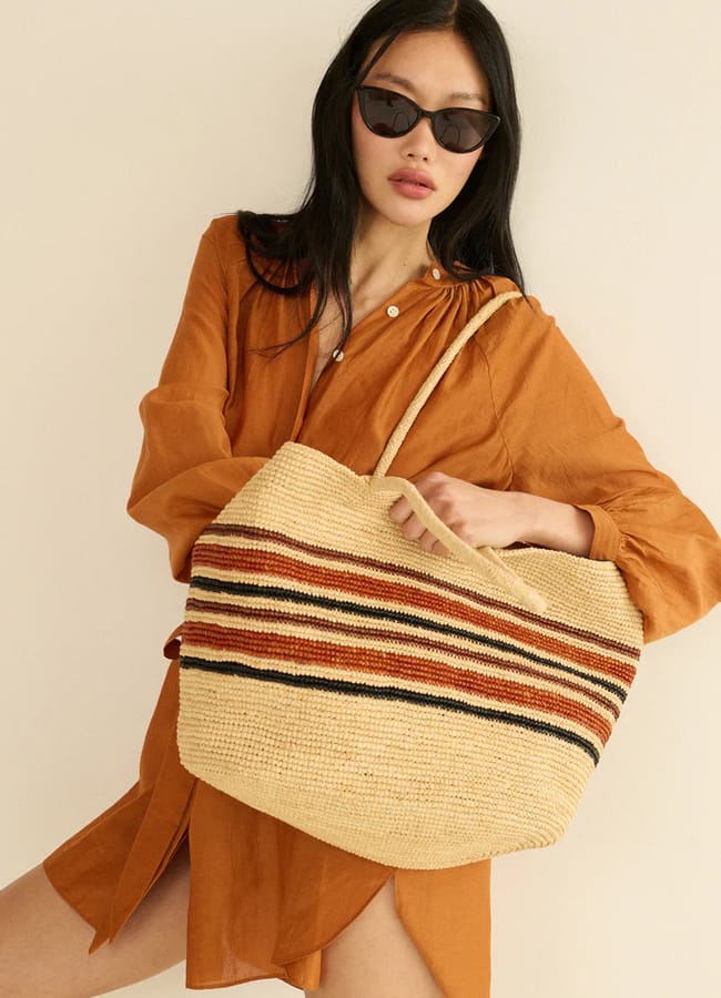 A woman wearing an orange shirt dress and black sunglasses poses with a large woven straw basket bag featuring red and black horizontal stripes. She stands against a plain light-coloured background.