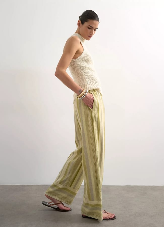 A person stands sideways against a plain white wall, wearing a sleeveless textured cream top, loose high street linen yellow and beige striped trousers, black sandals, and a bracelet, with hair tied back.