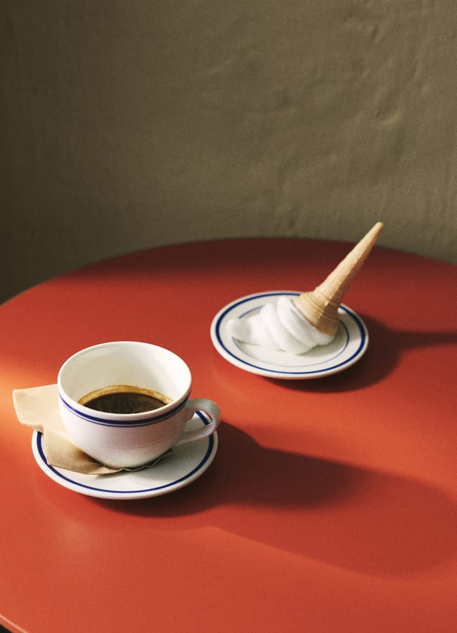 A cup of black coffee with a serviette sits on a saucer on a red table. Nearby, a melting ice cream cone is placed upside down on a small plate. The background wall is beige.