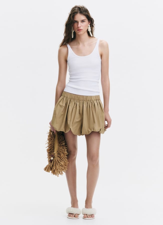 A woman stands against a white background, wearing a white vest and tan bubble shorts. She holds a brown woven clutch, pairs the look with white sandals, and lets her loose hair frame her neutral expression.