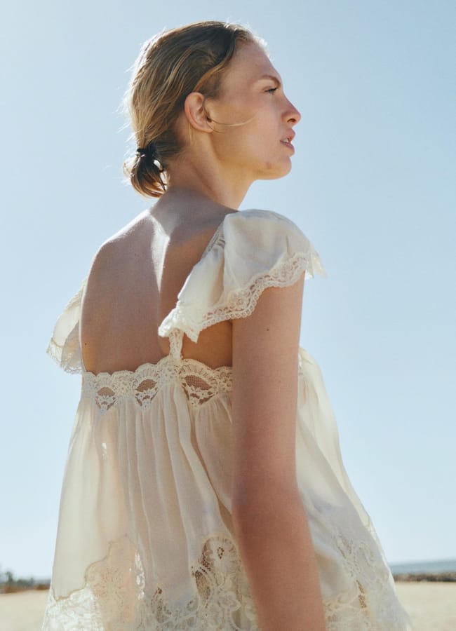 A woman with light brown hair in a low bun wears a cream-coloured, lace-trimmed summer top and looks into the distance outdoors on a sunny day, with a clear blue sky in the background.