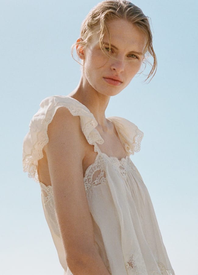 A woman with fair hair and delicate features stands outdoors under a clear sky, wearing a sleeveless, frilled white dress with lace details. She gazes softly at the camera, sunlight illuminating her face—an image as thoughtful as unique Fathers’ Day gifts 2026.
