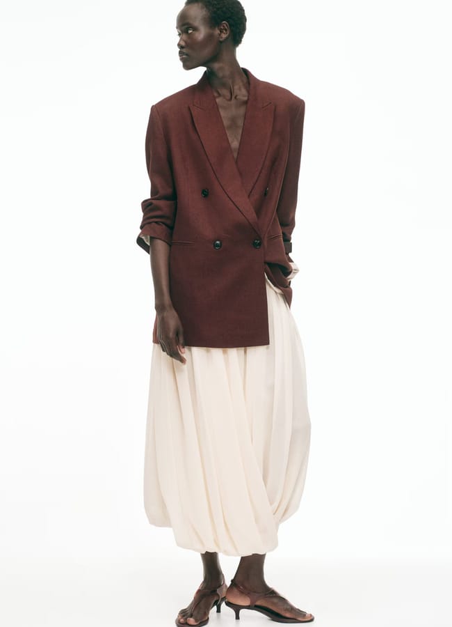 A person stands against a white background wearing a maroon double-breasted blazer, a loose high street linen skirt, and brown heeled sandals. They have short hair and their head is turned to the side.