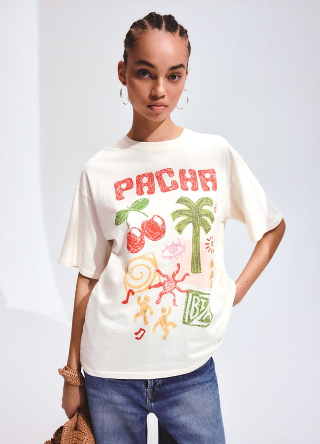 A woman with braided hair wears hoop earrings, a cream-coloured t-shirt with colourful fruit prints and the word PACHA, and blue jeans, standing against a plain, light background.