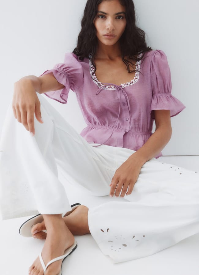 A woman with long dark hair sits against a white background, wearing a lavender short-sleeve summer top with embroidered trim, white wide-leg trousers with cut-out details, and white flip-flops.
