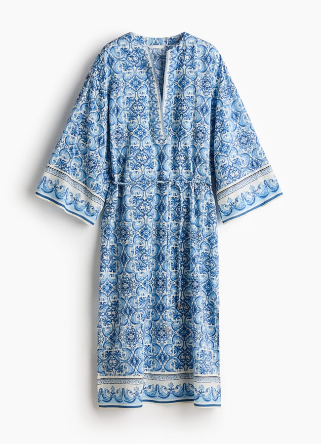 A loose, long-sleeved dress with a blue and white ornate pattern, featuring a V-neckline and decorative borders on the sleeves and hem. The dress has a relaxed, comfortable fit.