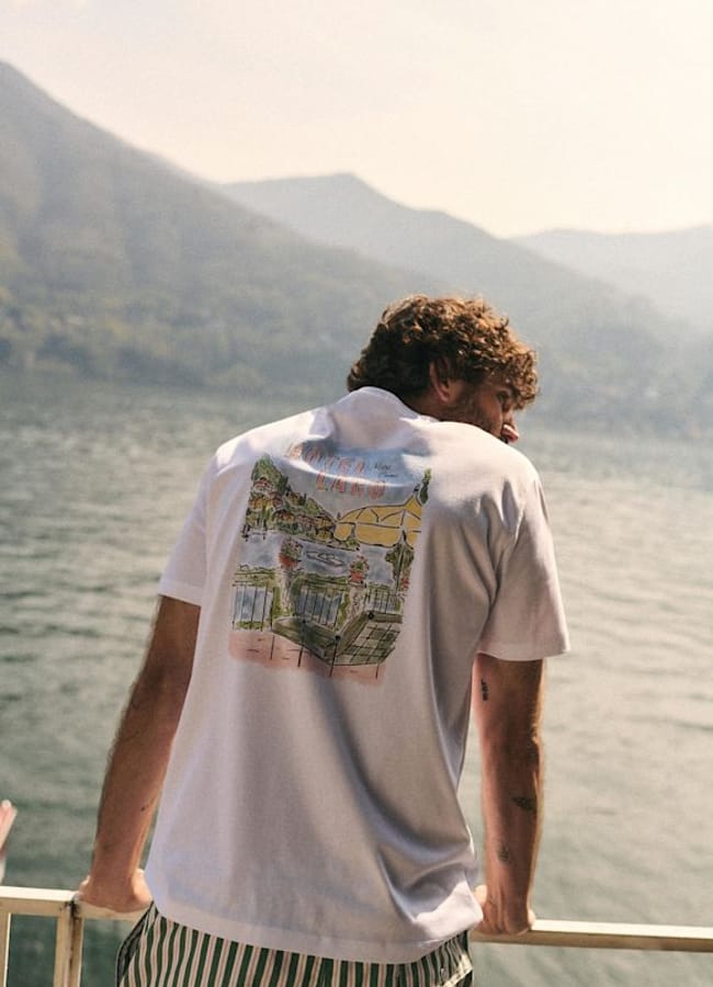 A man with curly hair leans on a railing, facing a lake surrounded by mountains. Showcasing men's holiday fashion, he wears a white T-shirt with a colourful illustrated back and green-striped shorts. The scene appears relaxed and summery.