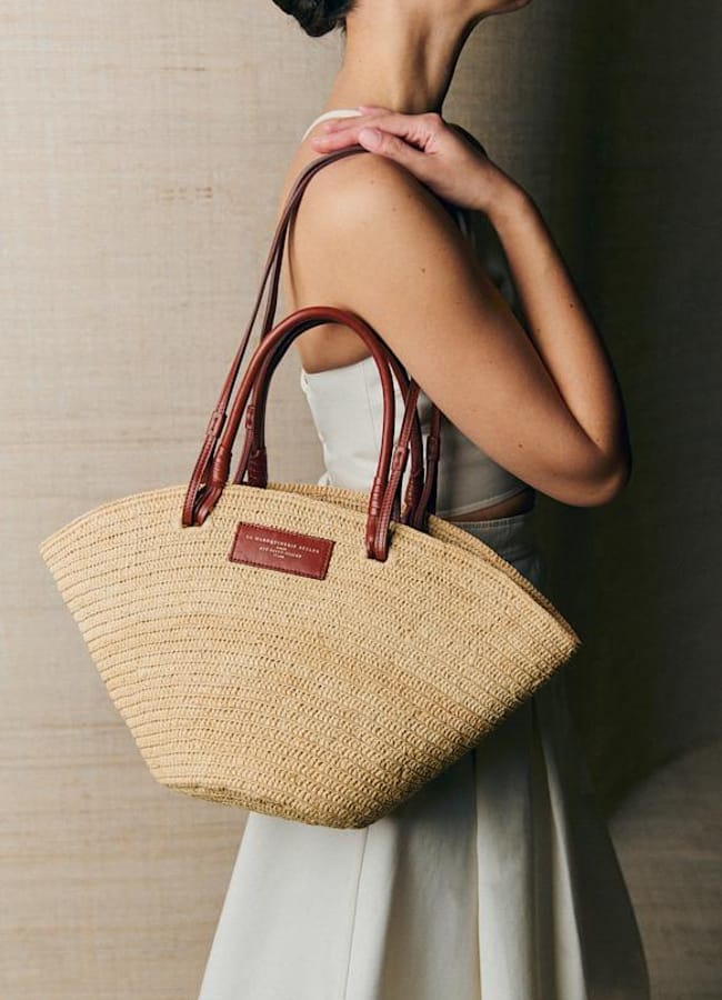 A woman in a sleeveless cream dress holds a woven straw basket bag with brown leather handles over her shoulder, standing against a neutral background.