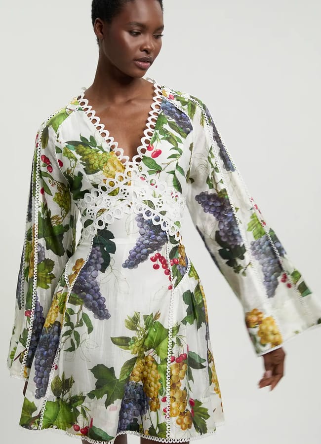 A woman wears a white dress with wide sleeves, decorated with vibrant fruit prints featuring grapes and currants. The dress has a deep V-neckline trimmed in white lace as she stands against a plain background.