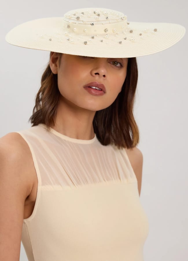 A woman wearing a sleeveless, sheer beige top and a wide-brimmed cream hat with decorative pearls—reminiscent of elegant fascinators—stands against a light background. The hat partially covers her eyes.