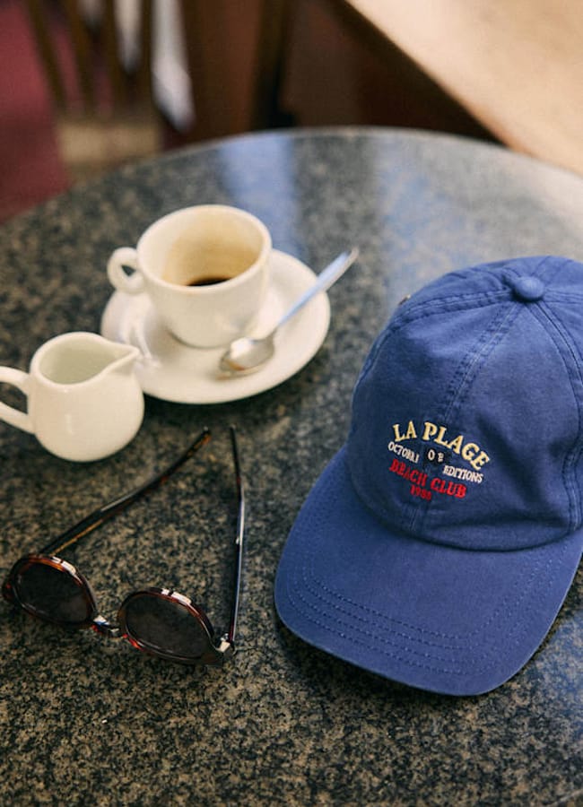 A blue baseball cap, sunglasses, a cup of coffee with a spoon, and a small jug of milk are arranged on a round, dark marble tabletop—capturing the essence of relaxed men's holiday fashion.