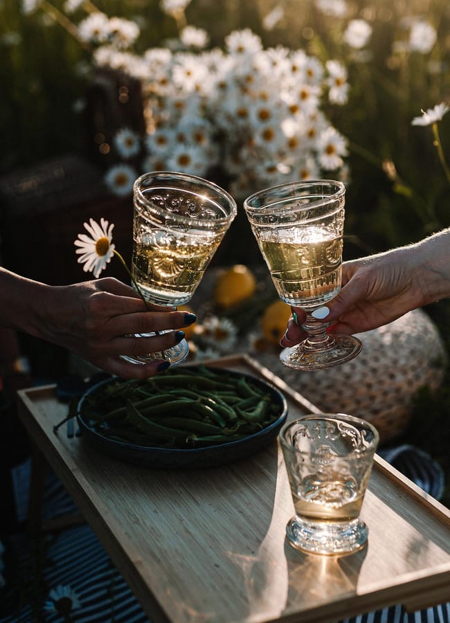 Two people clink glasses of white wine over a wooden tray with a bowl of green beans, set outdoors among blooming daisies in warm, golden sunlight.