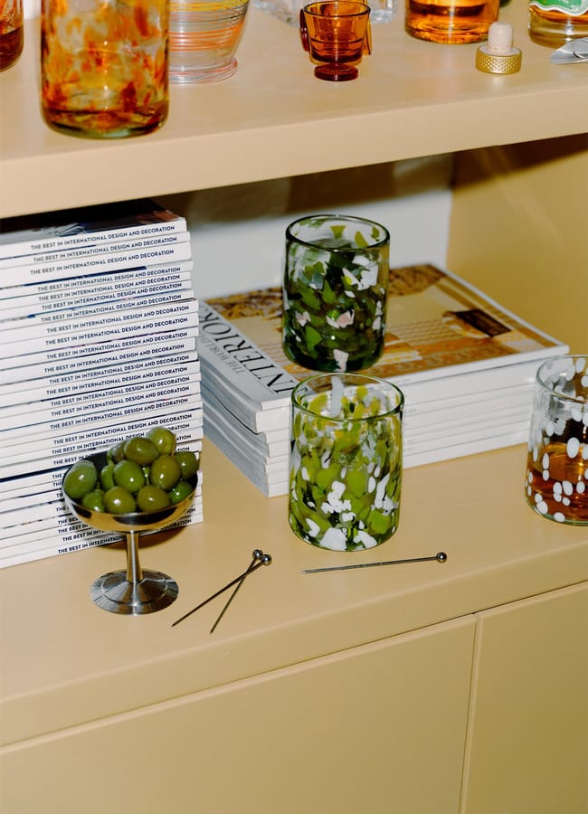 A beige shelf holding stacks of magazines, a metal dish of green olives, two decorative cocktail sticks, and colourful drinking glasses with green and white patterns.