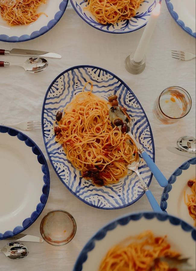 A serving platter with spaghetti and olives sits on a white tablecloth, surrounded by blue and white plates, glasses, cutlery, and a lit white candle.
