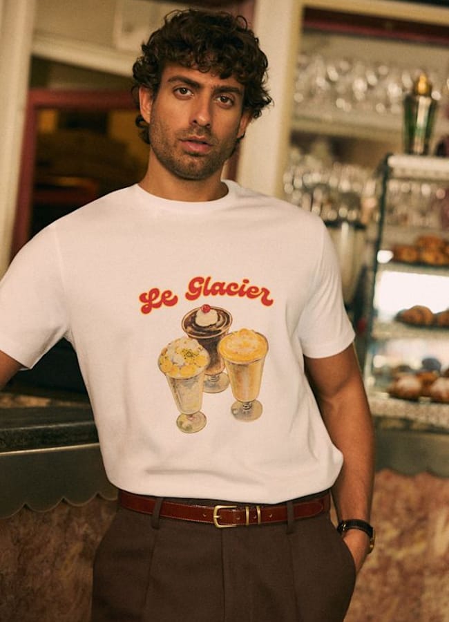 A man with curly hair and a trimmed beard stands indoors, embodying men's holiday fashion in a white Le Glacier T-shirt featuring three ice cream sundaes. He leans against a counter in a vintage-style café.