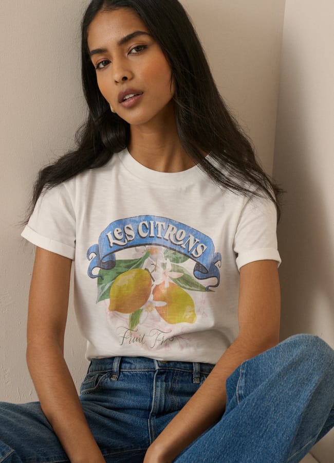 A woman with long dark hair wears a white T-shirt featuring vibrant fruit prints and the words Les Citrons, paired with blue jeans, as she sits against a light-coloured wall.