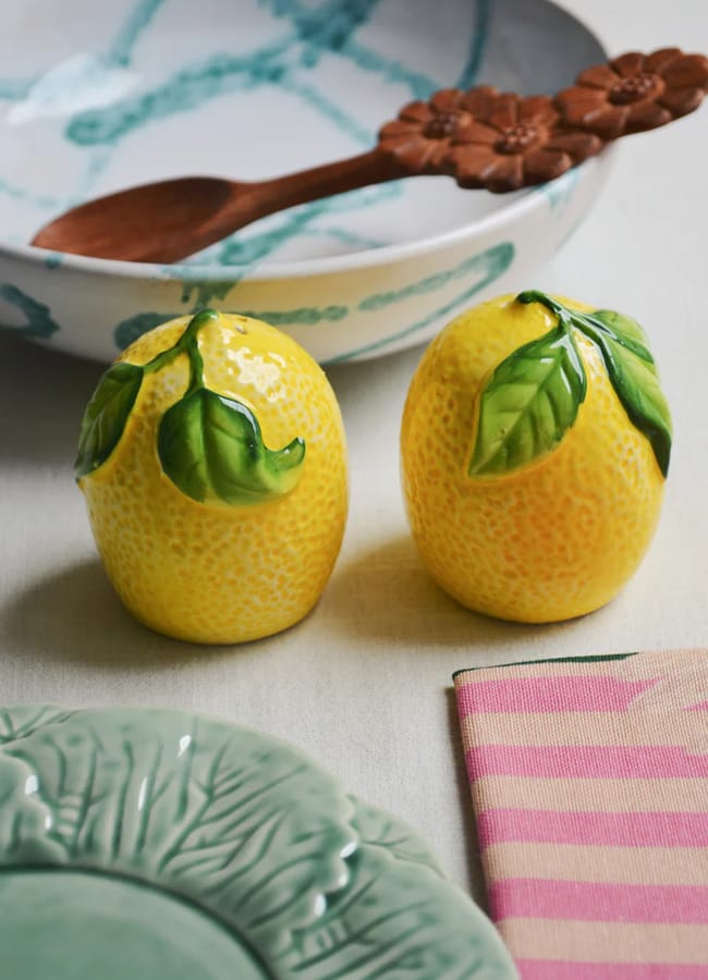 Two ceramic lemon-shaped shakers with green leaves sit on a table next to a green plate, a pink striped serviette, and a patterned bowl with a wooden spoon.