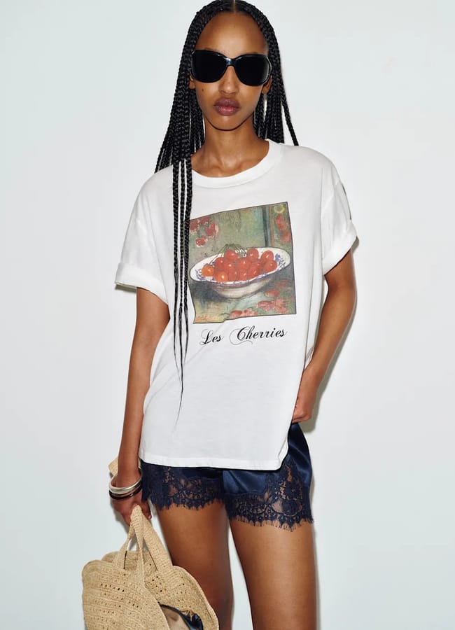 A woman with long plaits wears sunglasses, a white T-shirt featuring fruit prints—a bowl of cherries with the text Les Cherries—navy lace-trimmed shorts, and holds a woven straw bag, standing against a plain white background.