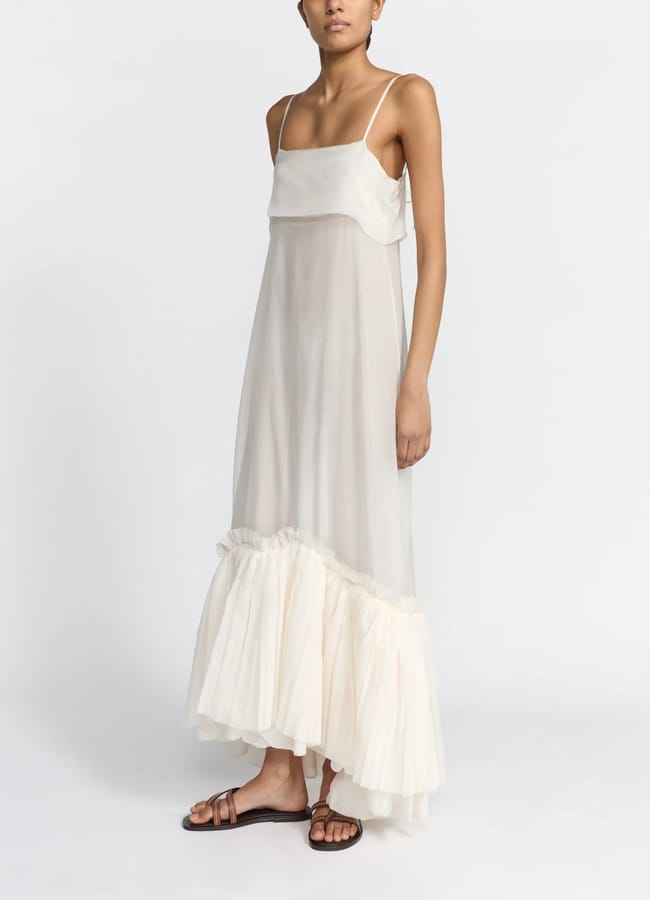 A person wears a floaty, sleeveless, off-white maxi dress with thin straps and a frilled hem, paired with brown flat sandals—an effortlessly chic look that could inspire unique Fathers' Day gifts 2026, standing against a plain white background.