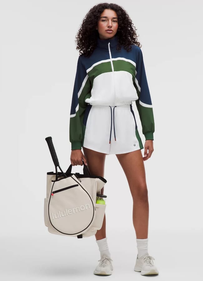A woman stands in athletic wear, including a long-sleeve zip-up top and shorts with navy, green, and white colour blocks—one of the cutest padel outfits. She holds a beige Lululemon sports bag with a tennis racquet and wears white trainers and socks.