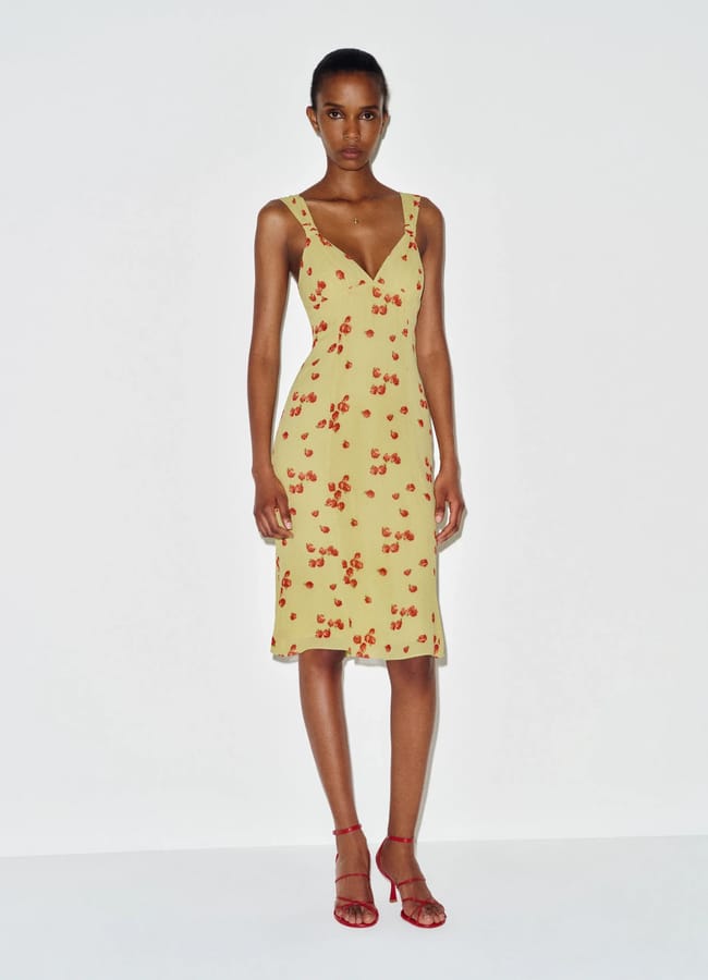A person stands against a plain white background wearing a yellow, knee-length dress with red fruit prints and red strappy high-heeled sandals.