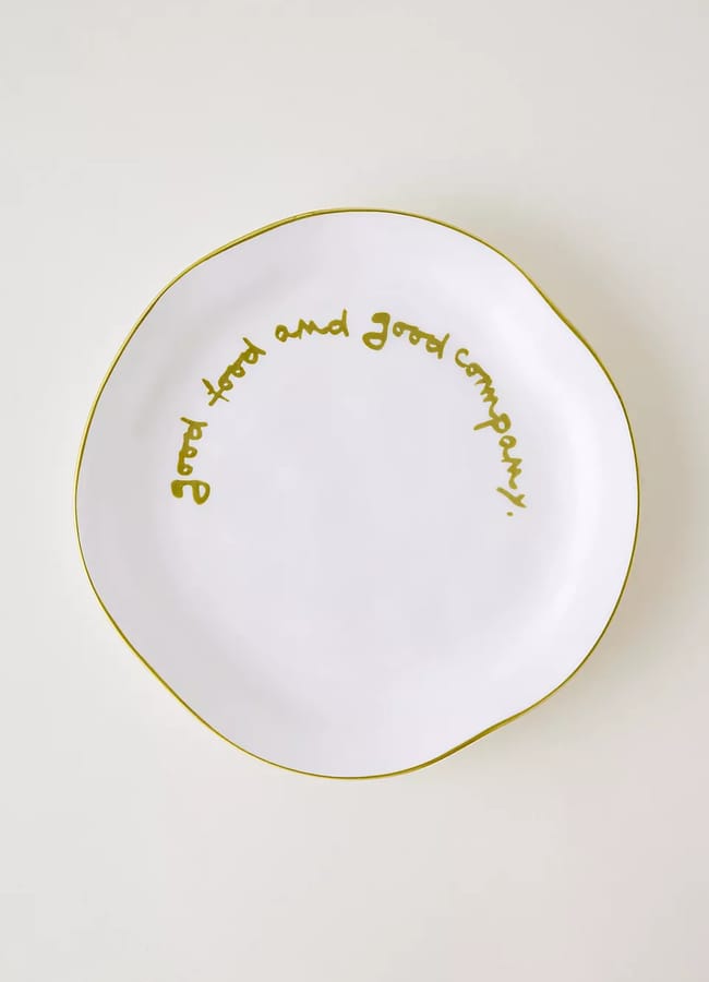 A white ceramic plate with a wavy edge outlined in gold, featuring the words “Good food and good company!” written in gold cursive along the rim.