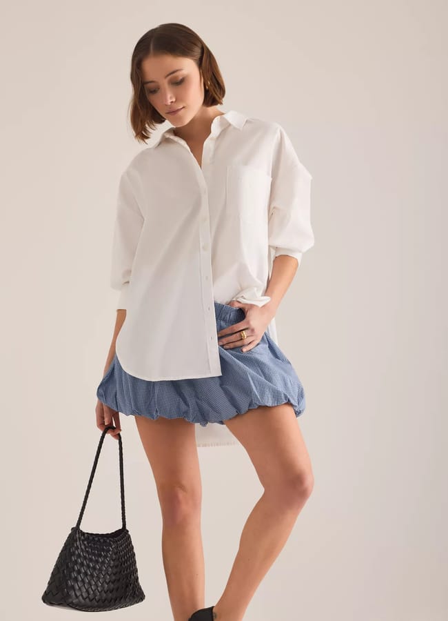 A woman stands against a plain background wearing an oversized white button-down shirt and short blue bubble shorts, holding a black woven handbag by her side.