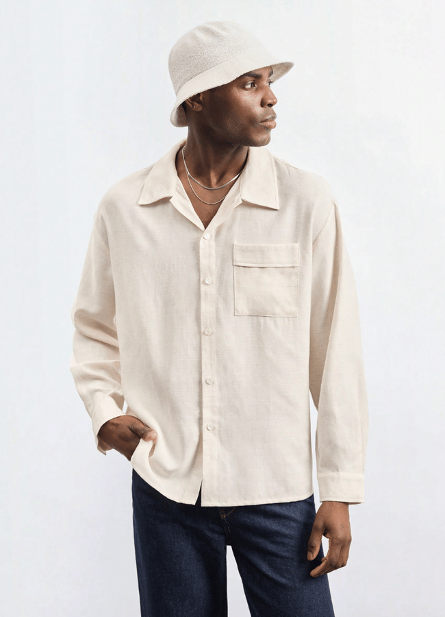 A man exemplifies men's holiday fashion in a light-coloured button-up shirt, dark jeans, a light bucket hat, and layered necklaces, standing with one hand in his pocket and gazing to the side against a plain background.