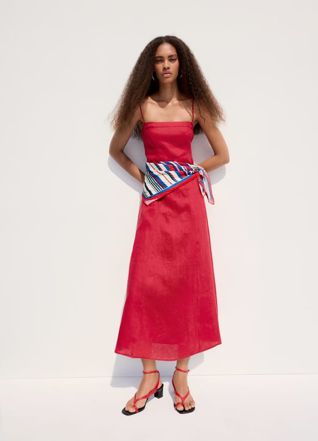 A woman stands against a white background wearing a sleeveless, high street linen midi dress in bright red, red strappy sandals, and a colourful striped scarf tied around her waist. She has long, voluminous hair and a confident expression.