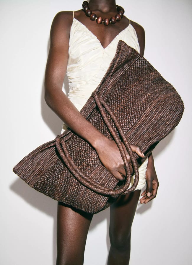 A person wearing a cream ruched dress and a chunky brown beaded necklace holds an oversized, woven dark brown basket bag against a plain background. The image is cropped from shoulders to mid-thigh.