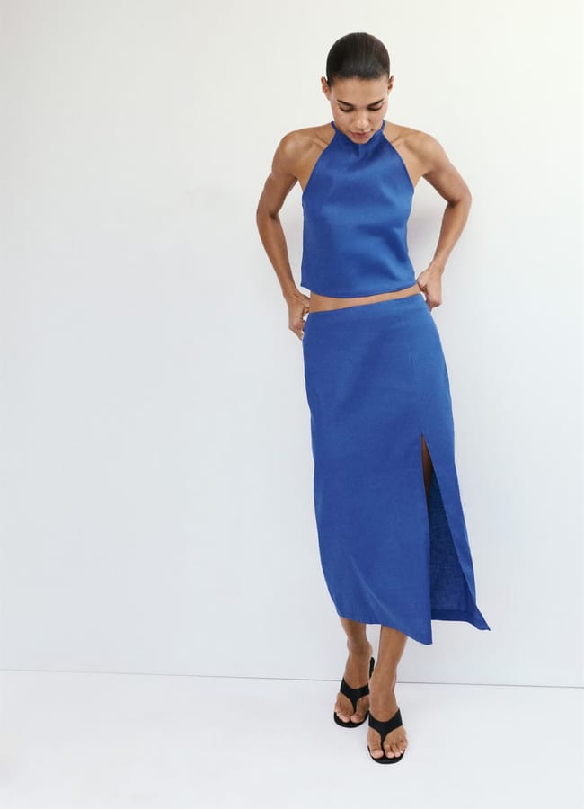 A person wearing a sleeveless, high-neck blue crop top and a matching high street linen midi skirt with a thigh-high slit, paired with black heeled sandals, stands against a plain white background.