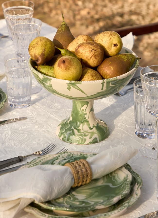 A green and white marbled pedestal bowl filled with pears sits on an elegantly set outdoor table with glassware, plates, cutlery, and a cloth serviette in a woven ring.