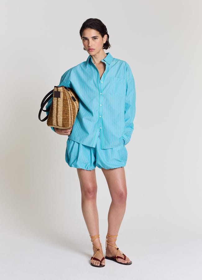 A woman stands against a plain background wearing a loose turquoise striped shirt and matching bubble shorts. She holds a straw tote bag and wears brown strappy sandals.