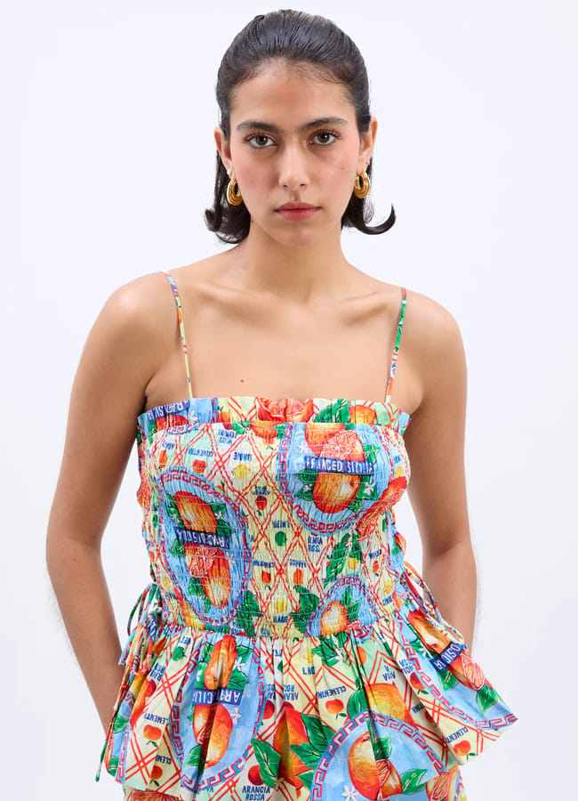 A woman with dark hair tied back wears gold hoop earrings and a sleeveless top featuring vibrant fruit prints with thin straps and a ruffled waist, standing against a plain white background.