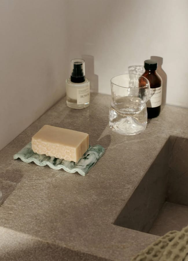A beige bar of soap on a ceramic dish rests on a stone worktop next to a glass, a pump bottle, and two brown bottles, reflecting affordable interiors in this minimalist bathroom setting.