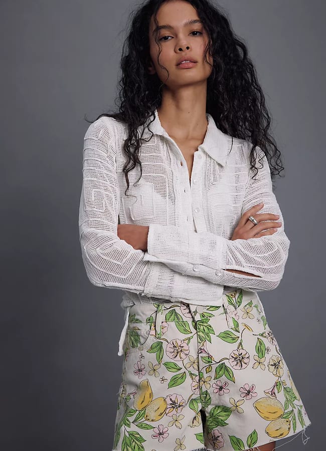 A woman stands with arms folded, wearing a sheer white long-sleeved blouse and high-waisted shorts featuring vibrant fruit prints and colourful florals, set against a plain grey background.